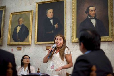 A woman speaks into the microphone and presents a case. Behind her, portraits of various famous men hang on the wall. 