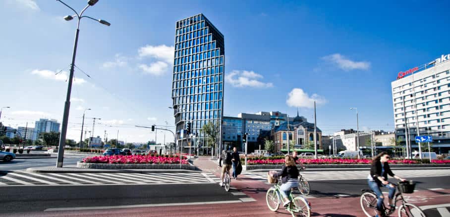 An image of Baltyk tower and the surrounding street intersection and people on bikes.. Baltyk tower is a large and modern office building with a glass front.