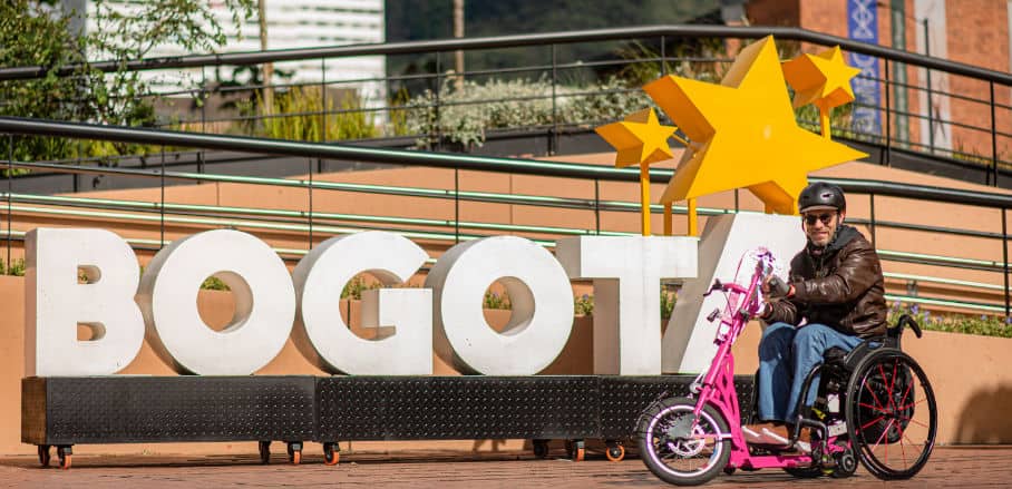 A man is riding a pink hand-bike, a bike for wheelchair users. He is standing in front of a large sign that reads "Bogotá".
