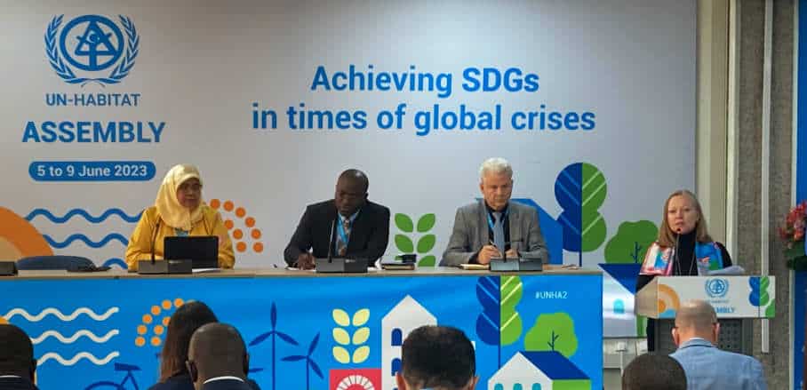 Leading figures of the UN Habitat Assembly sitting on a podium. In the back the theme of this year's assembly: Achieving SDGs in times of global crises.