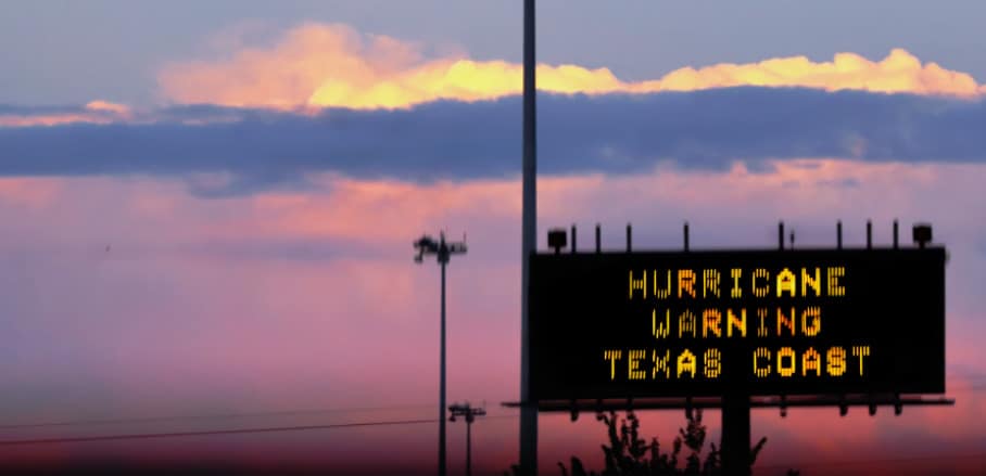 In front of a pink sky, a billboard says "Hurrican Warning Texas Coast"
