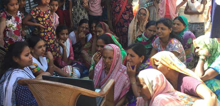 A crowd of Indian women jointly looks at a laptop screen that is placed on a chair outside.
