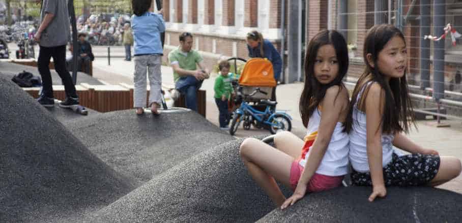 Children are sitting on a rubberised playsapce, with families in the background.