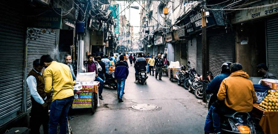 The picture shows a narrow street in India as well as people standing and on motorbikes going thorugh.